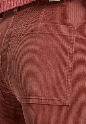 Materiał w kolorze rdzawym z korduroy, z pionowymi żebrami, dwiema tylnymi kieszeniami i teksturowaną powierzchnią. Zbliżenie na tkaninę i szczegóły projektu.