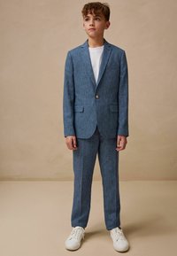 Completo due pezzi in tessuto azzurro chiaro con una trama sottile, composto da una giacca a un bottone e pantaloni leggermente aderenti, abbinato a sneaker bianche.