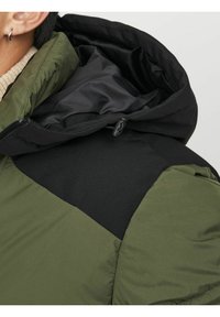 Giacca a vento verde e nera con texture trapuntata. Presenta un cappuccio nero, blocchi di colore a contrasto e un dettaglio con zip.