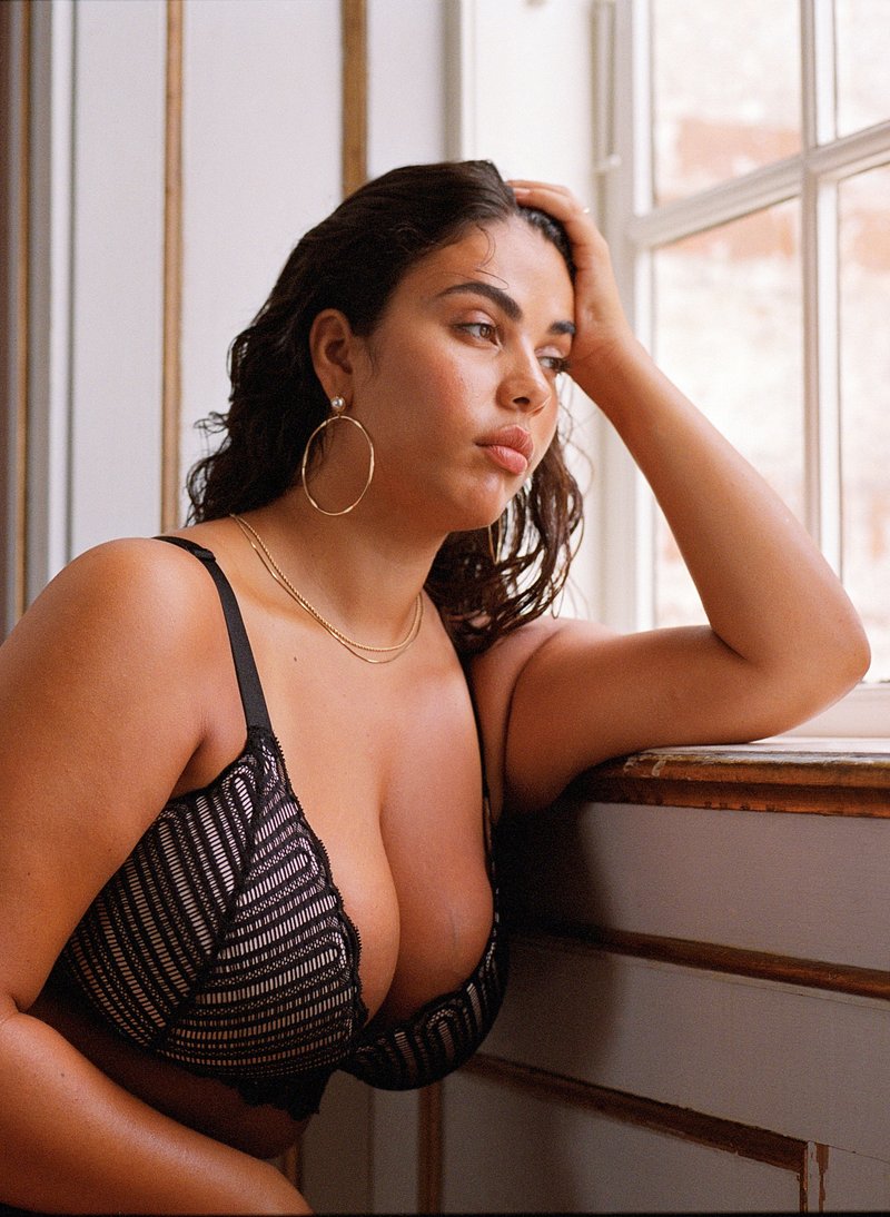 Black lace bralette with a striped pattern and scalloped edge. Model has hoop earrings and layered necklaces, seated near a window.