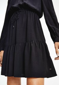 Schwarzes Kleid mit langen Ärmeln, verstellbarem Kordelzug in der Taille und geschichteter Saum. Weicher Stoff mit glatter Textur und Logo-Detail am Saum.