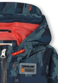 Veste pour enfants Timberland avec un motif camouflage bleu et vert, une fermeture éclair à l'avant, des accents orange et une doublure en mesh.