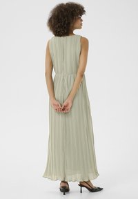 Plisserad, ärmlös maxikjol i ljusgrön, med nyckelhålsrygg och knappstängning samt svävande nederkant, parat med svarta klackar.