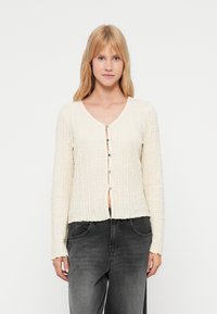 Cardigan crème avec un design texturé en tricot. Doté d'un col en V et d'une fermeture à boutons sur le devant. Manches longues avec une coupe décontractée.