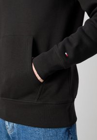 Main dans la poche d'un sweat-shirt noir avec un petit logo rouge, blanc et bleu sur la manche, porté avec un jean bleu.