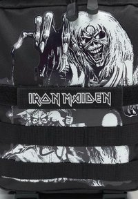 Čierny batoh s bielymi grafickými ilustráciami lebiek a postáv. Obsahuje logo "IRON MAIDEN" v tučnom písme na popruhu.