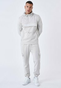 Lichtgrijze trainingsoutfit met een ritsachtige hoodie en een opdruk aan de voorkant, en elastische manchetten. De broek heeft cargozakken en een elastische tailleband.