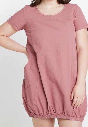 Femme portant une robe rose poudré à manches courtes avec poches latérales et ourlet froncé, debout, une main sur la hanche et l'autre le long du corps.