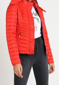 Femme portant une veste matelassée rouge vif sur un t-shirt graphique blanc et un jean taille haute noir, debout bras détendus.