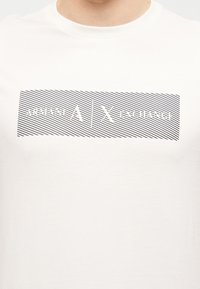 Λευκό μπλουζάκι με οριζόντιες μαύρες γραμμές και την επιγραφή «ARMANI | EXCHANGE» σε λευκό κείμενο μέσα σε ορθογώνιο πλαίσιο στο στήθος.