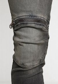 Pantalones de mezclilla gris con un bolsillo con cremallera en la rodilla, que presentan una textura ligeramente deslavada y un diseño ajustado.