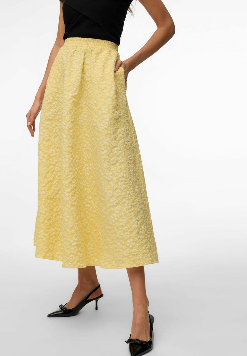 Femme portant une jupe midi texturée jaune à motifs floraux, un haut noir sans manches, et des escarpins noirs à bout pointu avec des brides arrière et des nœuds.