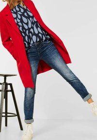 Femme portant un manteau rouge, un haut abstrait bleu marine et bleu clair, un jean retroussé et des baskets blanches, se tenant avec une jambe levée à côté d'un tabouret noir.