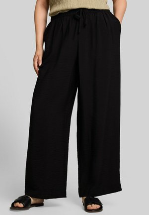 Femme portant un pantalon noir à jambes larges avec une taille à cordon et des sandales plates noires ouvertes à l'avant, debout les mains dans les poches.
