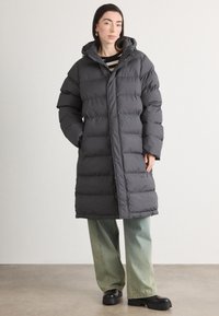 Abrigo acolchado en gris oscuro, con capucha, diseño acolchado, corte oversize y cierre frontal. Combinado con jeans verdes de pierna ancha y botas negras.