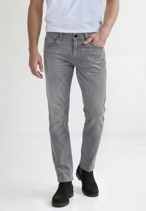 Mand iført grå slim-fit jeans med den ene hånd i lommen, kombineret med sorte sko og en hvid skjorte, stående foran en ensfarvet baggrund.