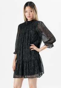 Jovem mulher com cabelo longo, vestindo um vestido preto transparente com padrão de zebra e mangas bufantes, de pé com uma mão na cintura.