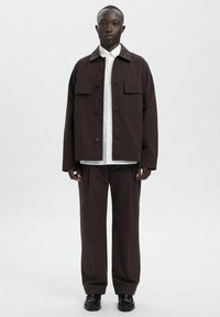 Tumepruun jakk kahe rindtaskuga, valge särk all, tumepruunad püksid ja mustad kingad; siledapinnaline, lõdvestunud lõige.