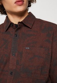 Brun flanellskjorta med svart texturerad mönster, knappad krage, bröstficka och logomärke. Knapparna är mörka och framträdande.