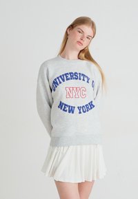 Jovem mulher vestindo um moletom cinza da "Universidade de Nova Iorque NYC" e uma saia plissada branca, de pé contra um fundo branco liso.