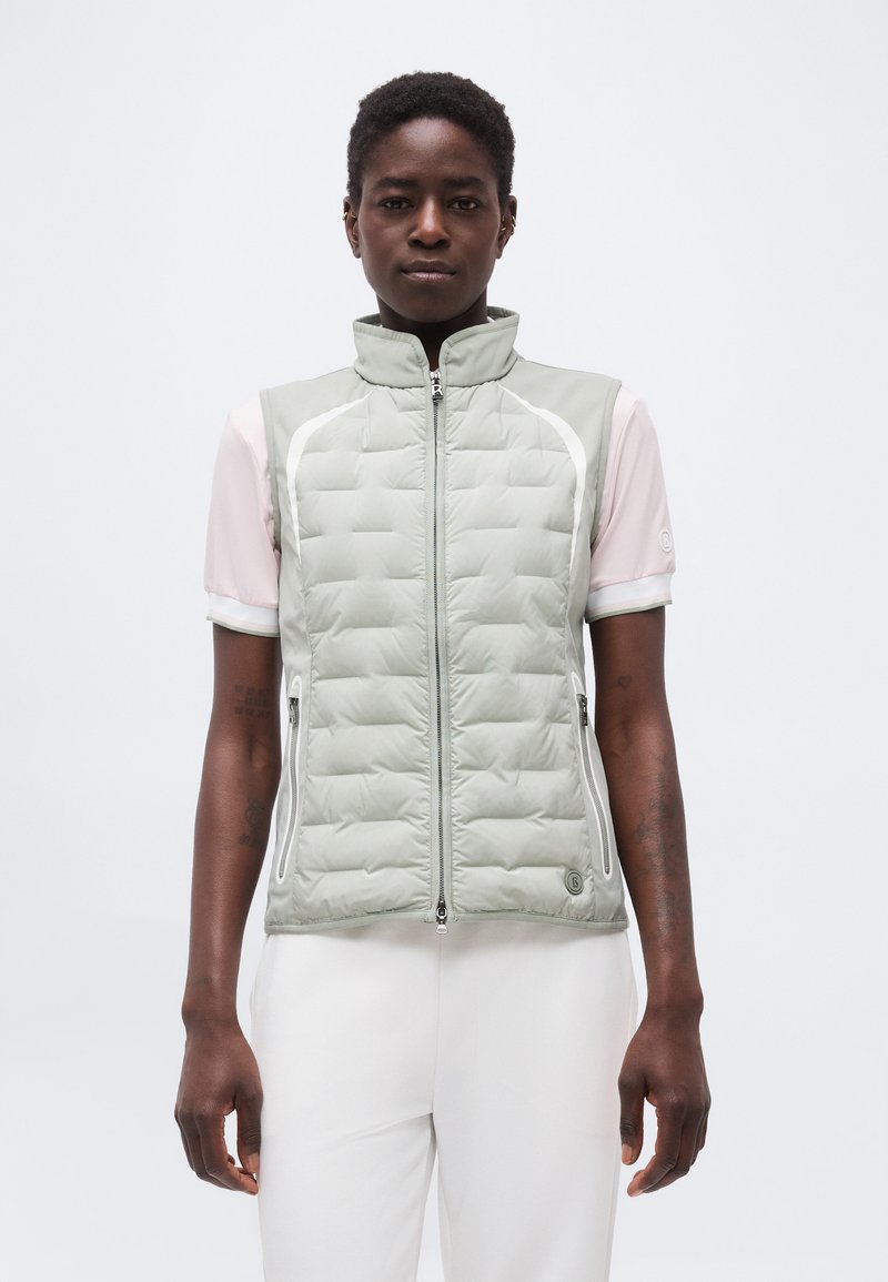 Γυναίκα με κοντά μαλλιά που φοράει ανοιχτό γκρι γιλέκο καπιτονέ πάνω από κοντομάνικο απαλό ροζ πουκάμισο και λευκό παντελόνι, όρθια μπροστά σε λευκό φόντο.