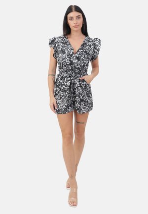 Kobieta z długimi ciemnymi włosami, ubrana w czarno-biały, kwiatowy kombinezon z falbankami na rękawach, ręce w kieszeniach i przezroczyste paski na obcasach.