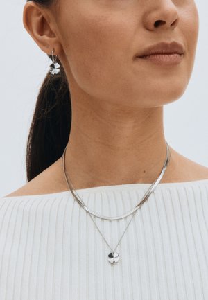 Silver necklace with two chains and a four-leaf clover pendant; matching silver earrings. Model wears a ribbed white top.