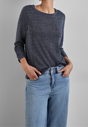 Personne portant un haut manches longues bleu foncé et un jean taille haute bleu clair, les mains dans les poches, devant un fond gris uni.