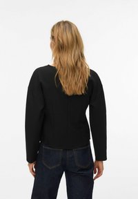 Juodos spalvos, aptemptas viršus su ilgomis rankovėmis, apvalia iškirpte ir struktūruotais pečiais. Turi lygią tekstūrą ir be siūlių nugaros dizainą.