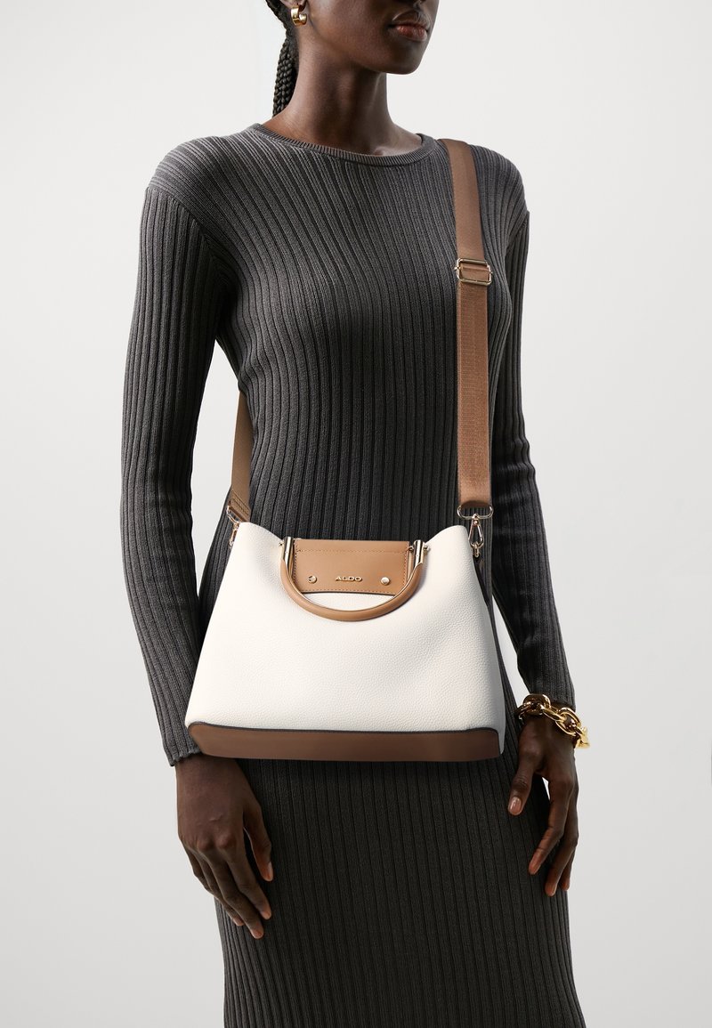 Femme portant une robe grise côtelée à manches longues, tenant un sac bandoulière blanc et marron avec des ferrures dorées.