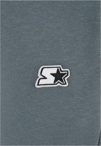 Siv gray material nežne teksturo krasijo vezenega logotipa v beli in črni barvi, ki prikazuje stilizirano črko 'S' in zvezdo.