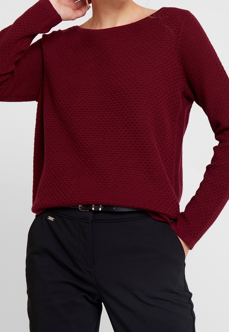 Femme portant un haut bordeaux texturé à manches longues rentré dans un pantalon noir avec une ceinture fine noire, la main posée près du cou et dans la poche.