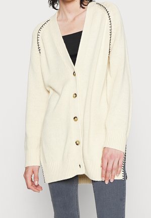 Cardigan crème oversize avec coutures noires aux épaules, fermeture par boutons, porté sur un haut noir et un pantalon gris, montré du cou à la mi-cuisse.