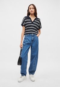 Μπλε σκούρο ριγέ πλεκτό τοπ με V-λαιμόκοψη και κοντά μανίκια, σε συνδυασμό με ψηλόμεσα, φαρδιά μπλε τζιν και λευκά αθλητικά παπούτσια, κρατώντας μια μαύρη τσάντα.