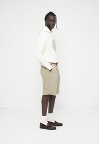 Μπεζ σορτς και κρεμ φούτερ με κουκούλα και κυκλικό γραφικό σχέδιο, συνδυασμένα με λευκές κάλτσες και γυαλιστερά μπουρντό loafers.