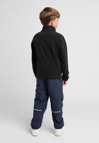 Pull en polaire noir avec un col haut, associé à un pantalon coupe-vent bleu marine doté de bandes réfléchissantes, complété par des baskets blanches.