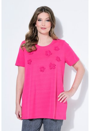 Kvinde i stor, lys pink kortærmet skjorte med blomsterapplikationer, stående med den ene hånd på hoften mod en hvid baggrund.