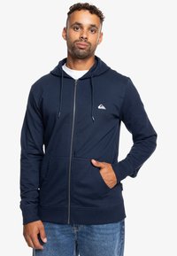 Námořnická mikina na zip s předními kapsami, nastavovací kapucí na šňůrku a malým bílým logem na levé hrudi. Měkká tkanina z bavlny a polyesteru.