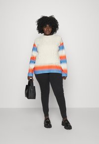 Suéter de malha com padrão de tranças em creme, com mangas longas em gradiente laranja e azul. Combinado com leggings pretas e sapatos de plataforma.