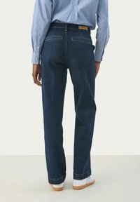 Jeans azul oscuro con un diseño de pierna recta, costuras visibles y dos bolsillos traseros, combinados con zapatillas blancas y una camisa de rayas.