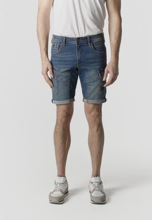 Mann trägt hochgekrempelte blaue Jeans-Cargoshorts, weiße Turnschuhe und ein schlichtes weißes T-Shirt und steht vor einem hellen Hintergrund.
