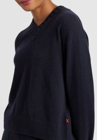 Pull en laine bleu marine avec un col en V, un col et un bas côtelés. Présente un petit logo rouge en forme de X en bas à gauche. Texture douce.