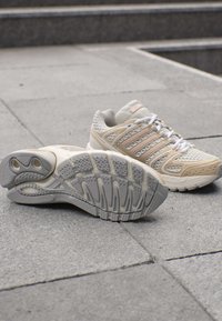 Paar beige und weiße Sportschuhe auf grauem Pflaster, ein Schuh zeigt eine strukturierte Sohle, der andere steht aufrecht mit gebundenen Schnürsenkeln.