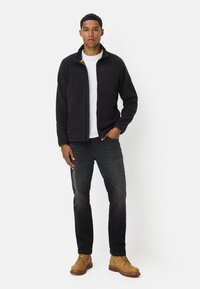 Zwarte fleecjas met ritssluitingen, gecombineerd met een witte t-shirt, donkere spijkerbroek en tan kleurige veterschoenen; staand tegen een witte achtergrond.