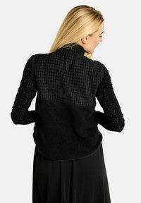 Melns vestes ar teksturētu aizmuguri un pūkainu apakšdaļu, kopā ar garām piedurknēm topu ar raibu rakstu.