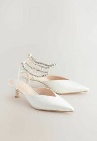 Valkoiset satiiniset slingback-kengät, joissa on kärkikärkinen muoto ja pieni korko. Koristeltu kirkkaista, nelikulmaisista strasseista koostuvalla ketjulla nilkkahihnassa.