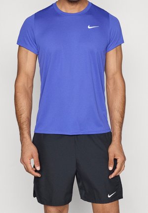Mann trägt ein blaues, kurzärmeliges Nike-Sportshirt und schwarze Nike-Shorts, steht vor einem einfarbigen Hintergrund.