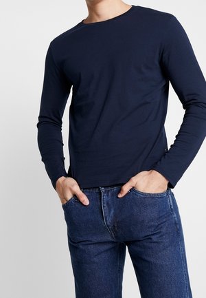 Hombre con camisa azul marino lisa de manga larga y vaqueros azules con las manos en los bolsillos frente a un fondo claro y sencillo.