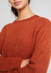 Femme portant de grandes boucles d'oreilles en anneau doré et un pull en tricot couleur rouille texturé avec un col à motif, les bras croisés.