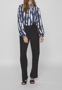 Blouse à manches longues à motifs bleu marine et blanc avec un col froncé, associée à un pantalon noir à jambe droite et des sandales à talons noires.